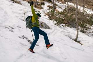 Eine Person in Winterkleidung mit Rucksack und Wanderstöcken rutscht auf einem schneebedeckten Hang aus. Im Hintergrund sind Bäume und Felsen zu sehen.