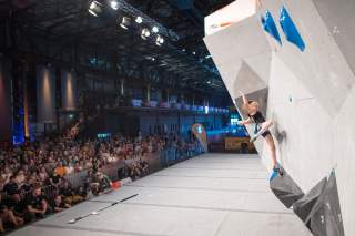 Frau klettert an einer Boulderwand bei einem Indoor-Kletterwettbewerb vor großem Publikum.