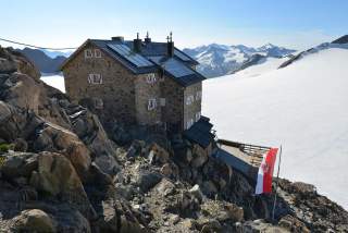 Steinernes Berghaus mit Solarpaneelen auf dem Dach steht auf felsigem Gelände neben einem schneebedeckten Gletscher, im Vordergrund weht eine rot-weiße Fahne mit Wappen.