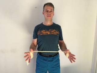 Ein Mann steht vor einer weißen Wand und hält mit beiden Händen ein gedehntes, beiges Gummiband, das um seine Handgelenke gewickelt ist. Er trägt ein blaues T-Shirt und blaue Shorts.