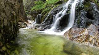 Kleiner Wasserfall fließt über moosbedeckte Felsen in einen klaren, grünen Gebirgsbach, umgeben von steilen Felswänden.