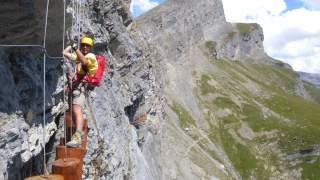 Ein Kletterer mit Helm und Sicherungsgurt überquert an einem Berg stehend eine Hängebrücke aus großen Holzscheiben. Im Hintergrund sind steile Felswände und ein bewachsener Berg zu sehen.