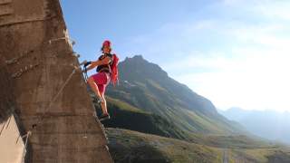 Frau mit Kletterausrüstung klettert an einer senkrechten Felswand, im Hintergrund sind Berge und ein blauer Himmel zu sehen.