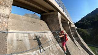 Eine Person mit Kletterausrüstung überquert an Seilen eine große Staumauer unter einer Brücke bei klarem Himmel. Im Hintergrund sind Berge und grünes Gelände sichtbar.