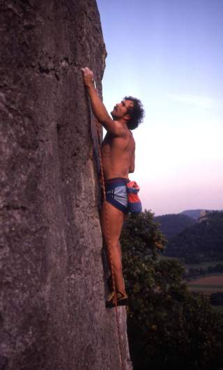 Ein shirtloser Mann klettert an einer steilen Felswand mit einfacher Kletterausrüstung bei Sonnenuntergang. Im Hintergrund sind bewaldete Hügel und ein weites Landschaftspanorama zu sehen.