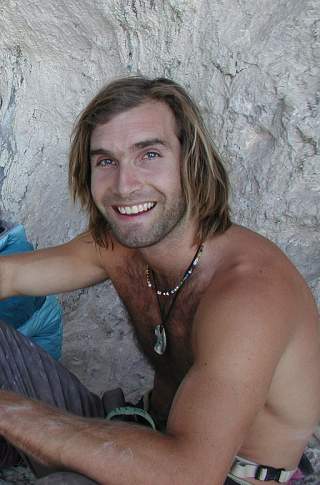 Lächelnder, muskulöser Mann mit schulterlangem Haar und Halskette sitzt shirtlos vor einer Felswand.