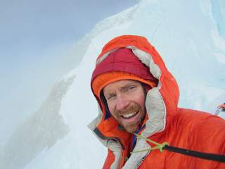 Nahaufnahme eines Mannes in orangefarbener Winterjacke und Mütze, der in einer verschneiten Berglandschaft lächelnd in die Kamera blickt.