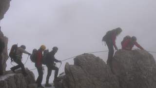 Fünf Bergsteiger mit Helmen und Rucksäcken sichern sich an einem Seil und klettern bei Nebel über felsiges, teils verschneites Gelände.