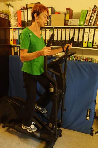 Ältere Frau in grünem Shirt trainiert auf einem Crosstrainer in einem Raum mit Aktenschränken und Bücherregalen.