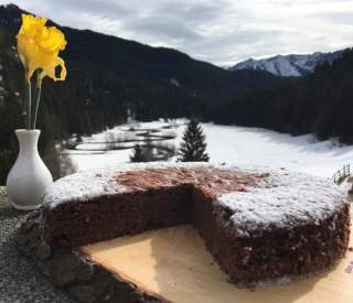 Schokoladenkuchen mit Puderzucker auf einem Holzbrett, im Hintergrund schneebedeckte Berge und eine weiße Vase mit zwei gelben Narzissen.