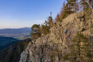 Eine Person klettert an einem felsigen Steilhang, umgeben von Bäumen und mit Blick auf ein Tal und entfernte Berge unter klarem Himmel.