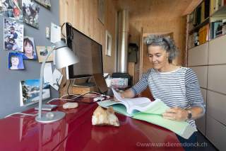 Eine Frau sitzt an einem roten Schreibtisch in einem Holzraum und liest in einem Buch, während ein Computerbildschirm, eine Lampe und Fotos an der Wand zu sehen sind.