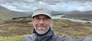 Ein Mann mit Brille, grauem Bart und Cap macht ein Selfie in einer hügeligen, kargen Landschaft mit einem Fluss oder See im Hintergrund unter einem bewölkten Himmel.
