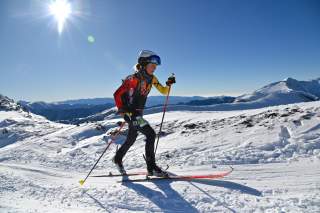 Person in bunter Skiausrüstung und Helm beim Skilanglauf in sonniger, verschneiter Berglandschaft.