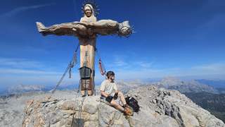 Ein Mann in Bergsteigerausrüstung sitzt auf einem Felsen neben einer großen geschnitzten Holzstatue eines gekreuzigten Jesus auf einem Berggipfel, mit weitem Blick auf die umliegenden Berglandschaften.