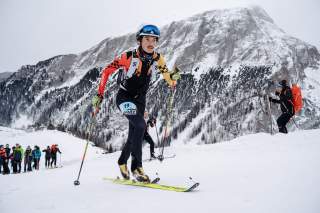 Skibergsteiger mit Rennanzug und Helm beim Aufstieg im Schnee vor einer schneebedeckten Bergkulisse, im Hintergrund weitere Teilnehmer und Zuschauer.