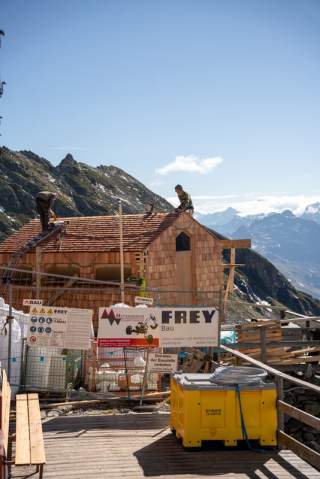 Zwei Arbeiter bauen an einem kleinen Holzhaus in den Bergen, umgeben von Bauzäunen, Schildern und Baumaterialien, im Hintergrund Alpenlandschaft.