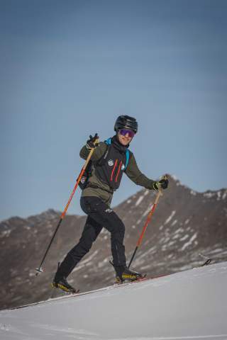 Skibergsteiger im Aufstieg, er lacht in die Kamera, im Hintergrund Berge