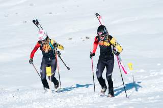 Eine Athletin und ein Athlet laufen nebeneinander die Piste hoch mit den Skiern auf dem Rücken. Beide tragen einen Rennanzug. Im Hintergrund sieht man die schneebedeckte Piste.