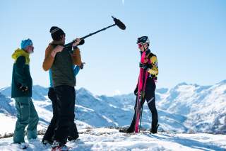 Auf der rechten Seite steht ein Athlet, ihm gegenüber drei Personen, die eine Kamera und ein Mikrofon halten. Im Hintergrund sind schneebedeckte Berge.