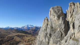 Ein Kletterer mit Helm und Kletterausrüstung erklimmt eine steile Felswand, im Hintergrund ist eine weite Berglandschaft unter klarem blauem Himmel zu sehen.