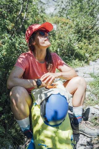 Frau mit rotem T-Shirt, roter Kappe und Sonnenbrille sitzt bei einer Wanderpause in der Sonne und isst eine Brezel.