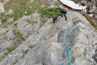 Zwei Bergsteiger mit Helmen klettern und sichern sich an einer Felswand, umgeben von felsigem Gelände und etwas Gras.