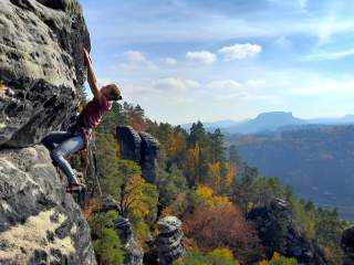 Eine Person klettert an einer steilen Felswand, im Hintergrund bewaldete Hügel und herbstlich gefärbte Bäume.