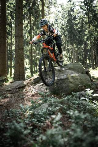Ein Kind mit Helm fährt mit einem Mountainbike über einen großen Felsen im Wald.