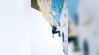 Ein junger Mann klettert an einer künstlichen Kletterwand im Freien, trägt Kletterausrüstung und blickt konzentriert nach oben. Im Hintergrund sind grüne Bäume und ein klarer Himmel zu sehen.