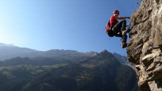 Ein Kletterer mit Helm und Rucksack klettert an einer steilen Felswand, im Hintergrund sind bewaldete Berge unter klarem blauem Himmel zu sehen.