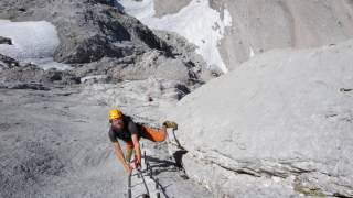 Ein Kletterer mit Helm und Kletterausrüstung steigt an Eisenstiften an einer steilen Felswand in den Bergen, im Hintergrund sind schneebedeckte Felsen zu sehen.