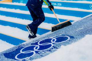 Eingeschneite Olympische Ringe, die jemand mit dem Besen frei fegt.