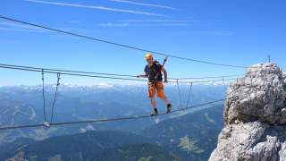 Ein Bergsteiger mit Helm und Sicherungsgurt überquert eine Drahtseilbrücke zwischen Felsen mit Blick auf eine bergige Landschaft unter blauem Himmel.