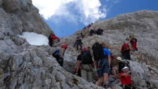 Eine Gruppe von Bergsteigern klettert an steilen Felsen entlang einer gesicherten Kletterroute unter blauem Himmel mit vereinzelten Wolken.