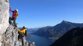 Zwei Personen klettern mit Ausrüstung an einer steilen Felswand, im Hintergrund sind ein See und bewaldete Berge unter blauem Himmel zu sehen.