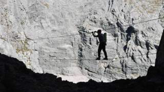 Silhouette einer Person mit Helm und Rucksack, die an einem Seilgesichert einen Drahtseilakt zwischen Felsen in einer Berglandschaft durchführt.