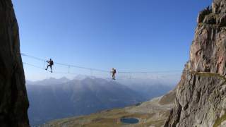 Zwei Personen überqueren eine schmale Hängebrücke zwischen steilen Felswänden mit Berglandschaft im Hintergrund, darunter ein kleiner See.