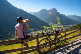 Radfahrer mit Helm sitzt auf einem Holzzaun, trinkt aus einer Flasche und blickt auf ein Tal mit Dorf und Bergen im Hintergrund, neben ihm steht ein Fahrrad.
