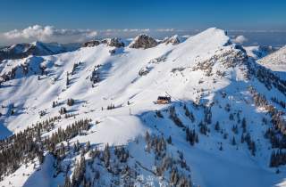 Verschneite Berglandschaft mit steilen Gipfeln, vereinzelten Bäumen und einer Berghütte in der Mitte.