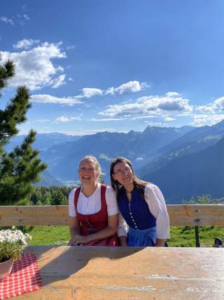 Zwei Frauen in traditioneller Tracht sitzen lachend auf einer Holzbank mit Bergpanorama im Hintergrund, bei sonnigem Wetter. Ein Tisch mit rotem Tuch und Blumentopf steht im Vordergrund.
