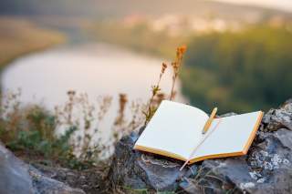 Ein leeres, kariertes Notizbuch mit Stift liegt auf einem Felsen, im Hintergrund ein unscharfer Fluss und grüne Landschaft.