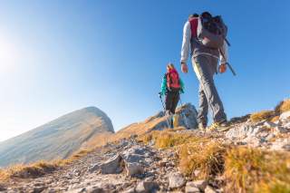 Zwei Wanderer mit Rucksäcken gehen bei sonnigem Wetter einen steinigen Bergpfad hinauf.