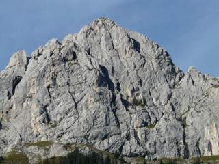 Felsiger Berggipfel mit einem Kreuz auf dem höchsten Punkt, darunter Wälder und Wiesen bei klarem Himmel.