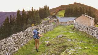 Ein Wanderer mit großem Rucksack und Wanderstöcken geht auf einem von niedrigen Steinmauern gesäumten Pfad zu einer kleinen Ansiedlung aus Steinhäusern in einer grünen, hügeligen Landschaft.