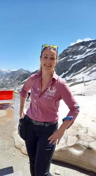 Frau in rotem kariertem Hemd steht lächelnd vor schneebedeckten Bergen bei klarem Himmel.