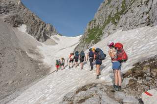 Gruppe von Wanderern mit Rucksäcken und Helmen steigt bei sonnigem Wetter über einen schneebedeckten Hang in einer felsigen Berglandschaft.