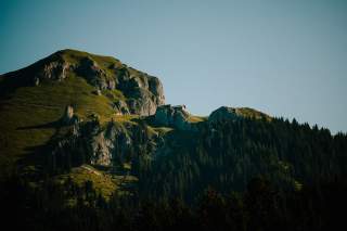 Berg mit grünen Wiesen und Felsen, darauf eine kleine Hütte, im Vordergrund dichter Nadelwald unter klarem Himmel.