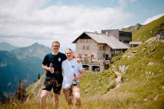 Zwei lächelnde Männer stehen auf einer Wiese in den Bergen, im Hintergrund ist eine Berghütte zu sehen.