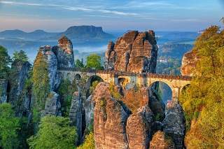Felsenformationen mit einer historischen Steinbrücke, umgeben von grünem und herbstlich gefärbtem Wald, unter blauem Himmel. Im Hintergrund sind weitere Berge und Täler zu sehen.